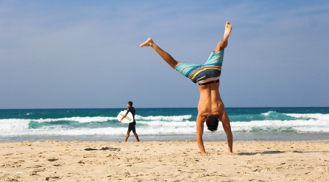 Man doing a handstand on the beach, surfer in background